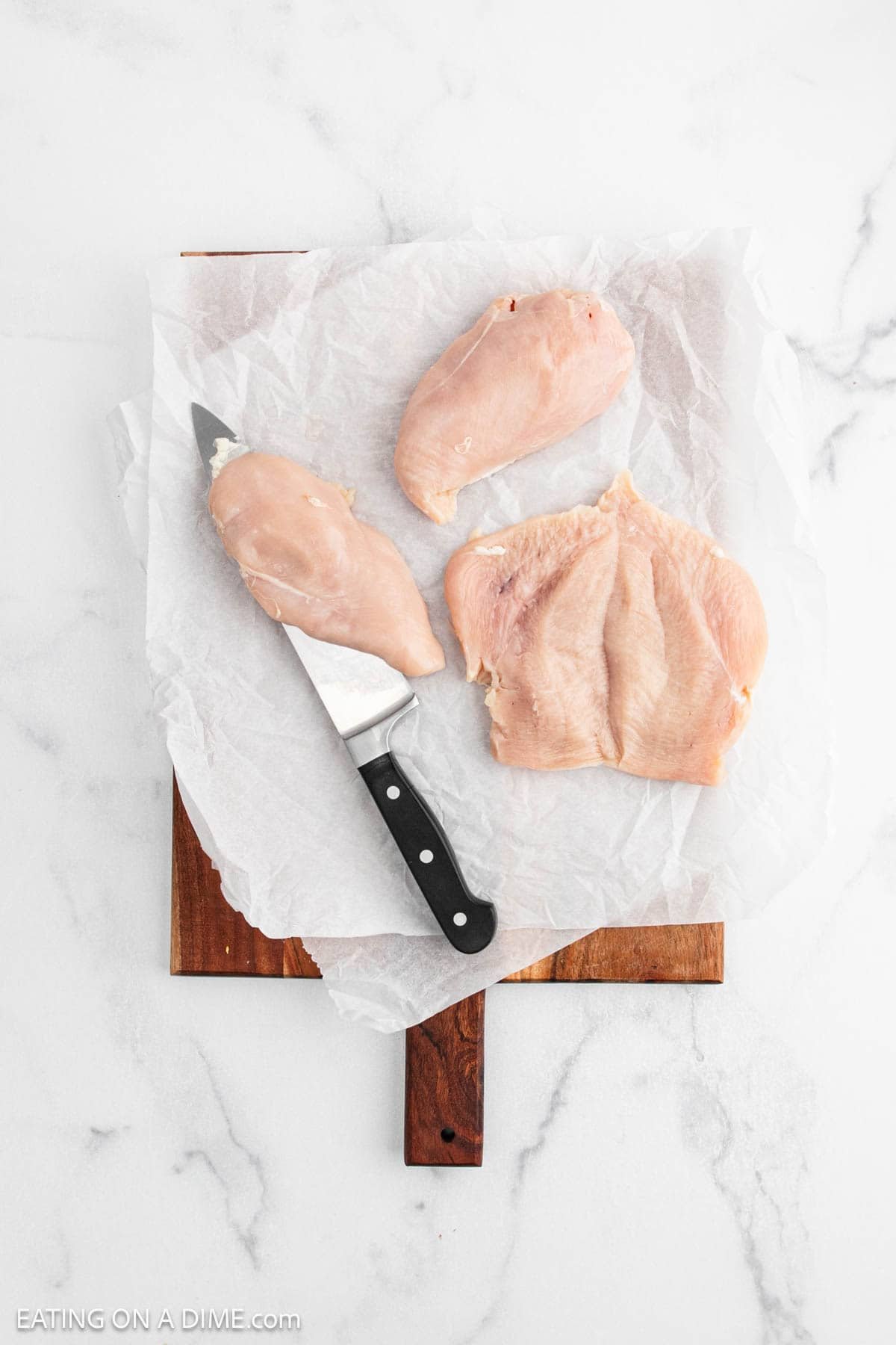 Raw chicken breasts, perfect for Chicken Fettuccine Alfredo, are shown—one whole and one sliced in half—on parchment paper atop a wooden cutting board. A kitchen knife lies beside them on a white marble surface.