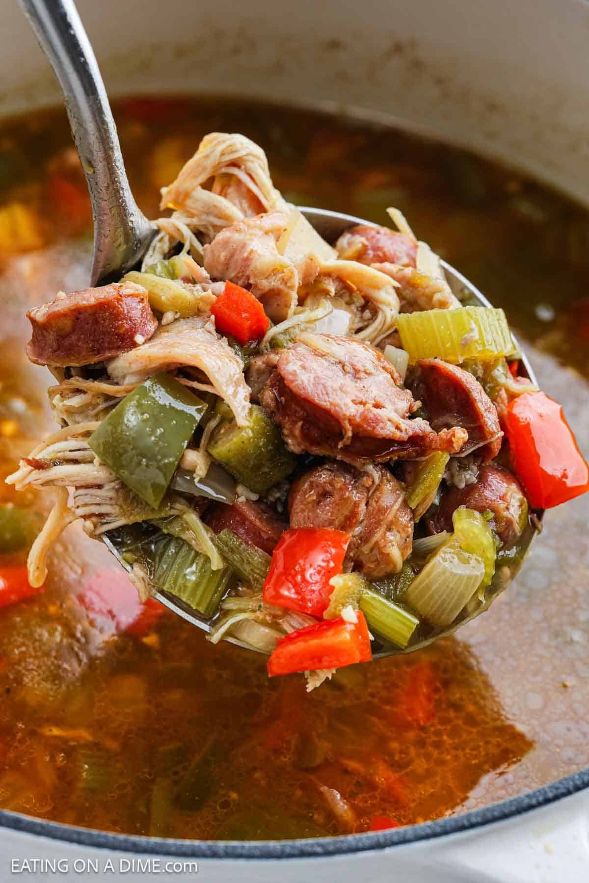 A close-up of a ladle lifting hearty Chicken Gumbo from a pot, showing chunks of sausage, shredded chicken, celery, and red and green bell peppers in a flavorful broth.