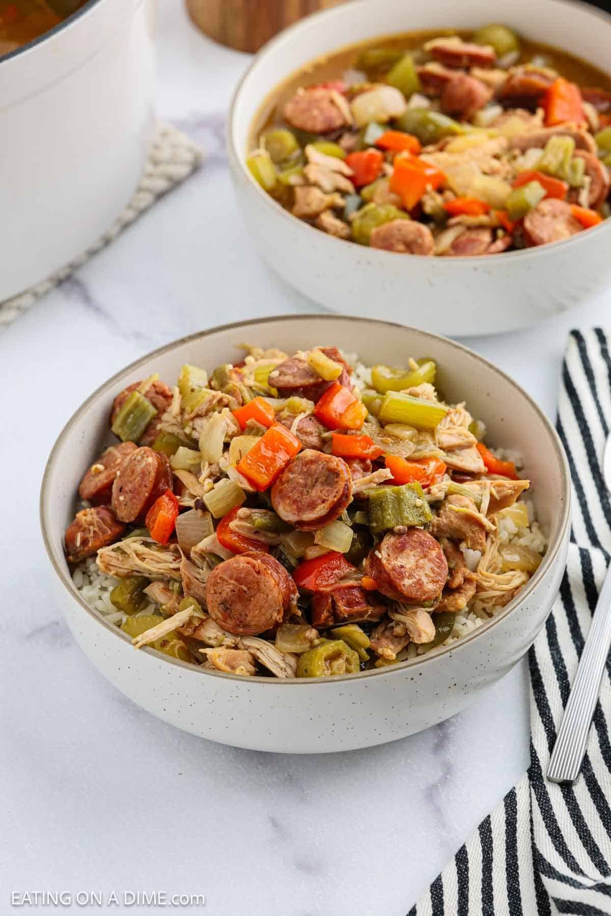 A bowl of chicken and sausage gumbo with rice, made from a classic Chicken Gumbo Recipe, is topped with chopped carrots, celery, and sausage slices. It sits on a white surface near a striped napkin and another bowl in the background.