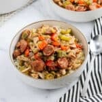 A bowl of rice topped with sliced sausage, shredded chicken, celery, red bell pepper, and onions—this dish brings Chicken Gumbo Recipe flavors to your table. Served with a spoon and striped napkin on a white surface.