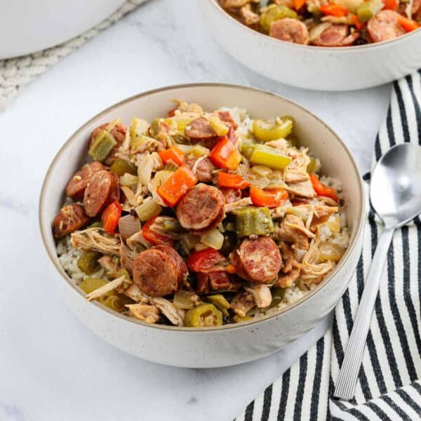 A bowl of rice topped with sliced sausage, shredded chicken, celery, red bell pepper, and onions—this dish brings Chicken Gumbo Recipe flavors to your table. Served with a spoon and striped napkin on a white surface.