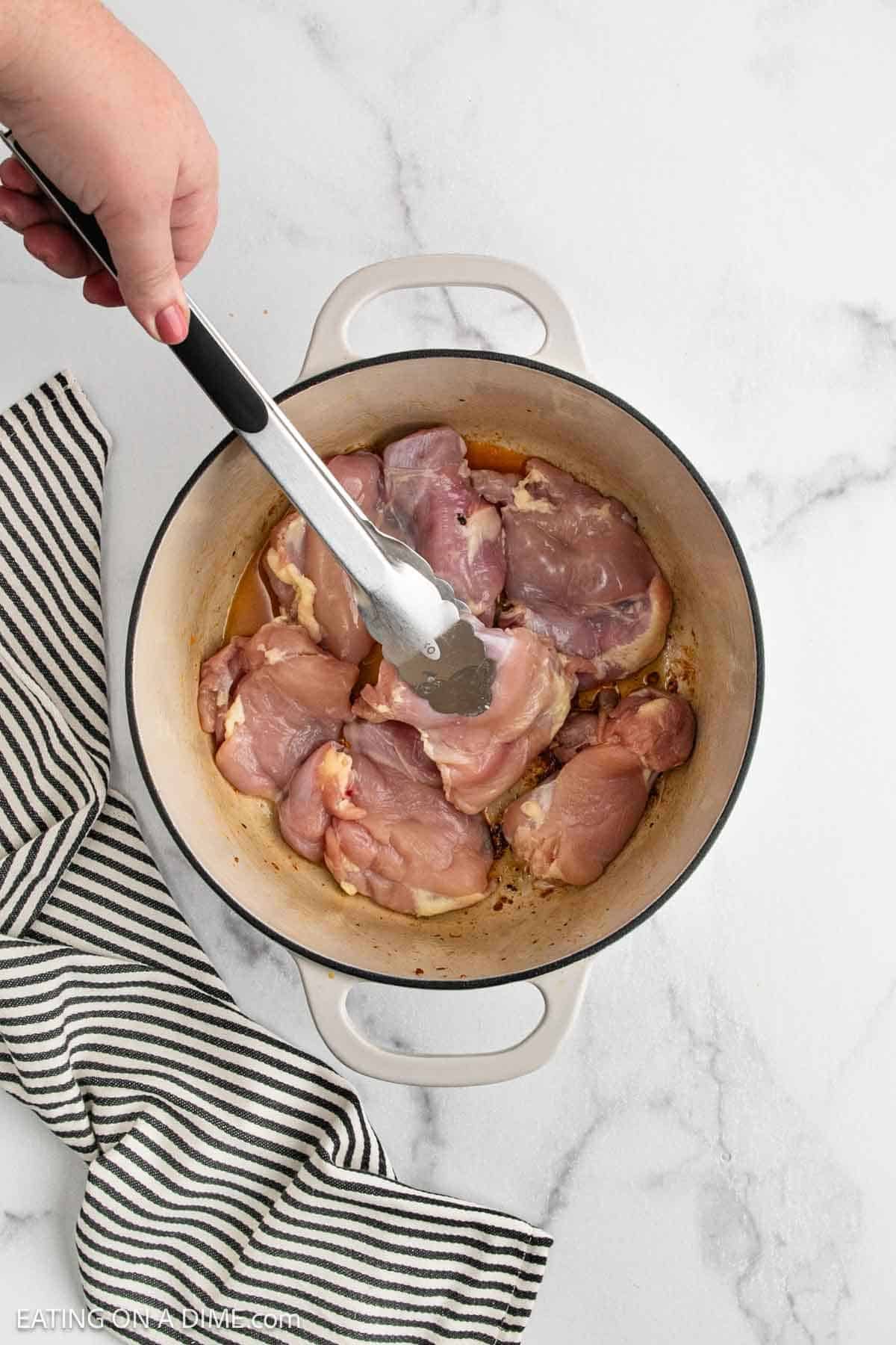 Hand using tongs to place raw chicken thighs into a white Dutch oven on a marble countertop, prepping ingredients for a delicious Chicken Gumbo Recipe, with a black and white striped towel nearby.
