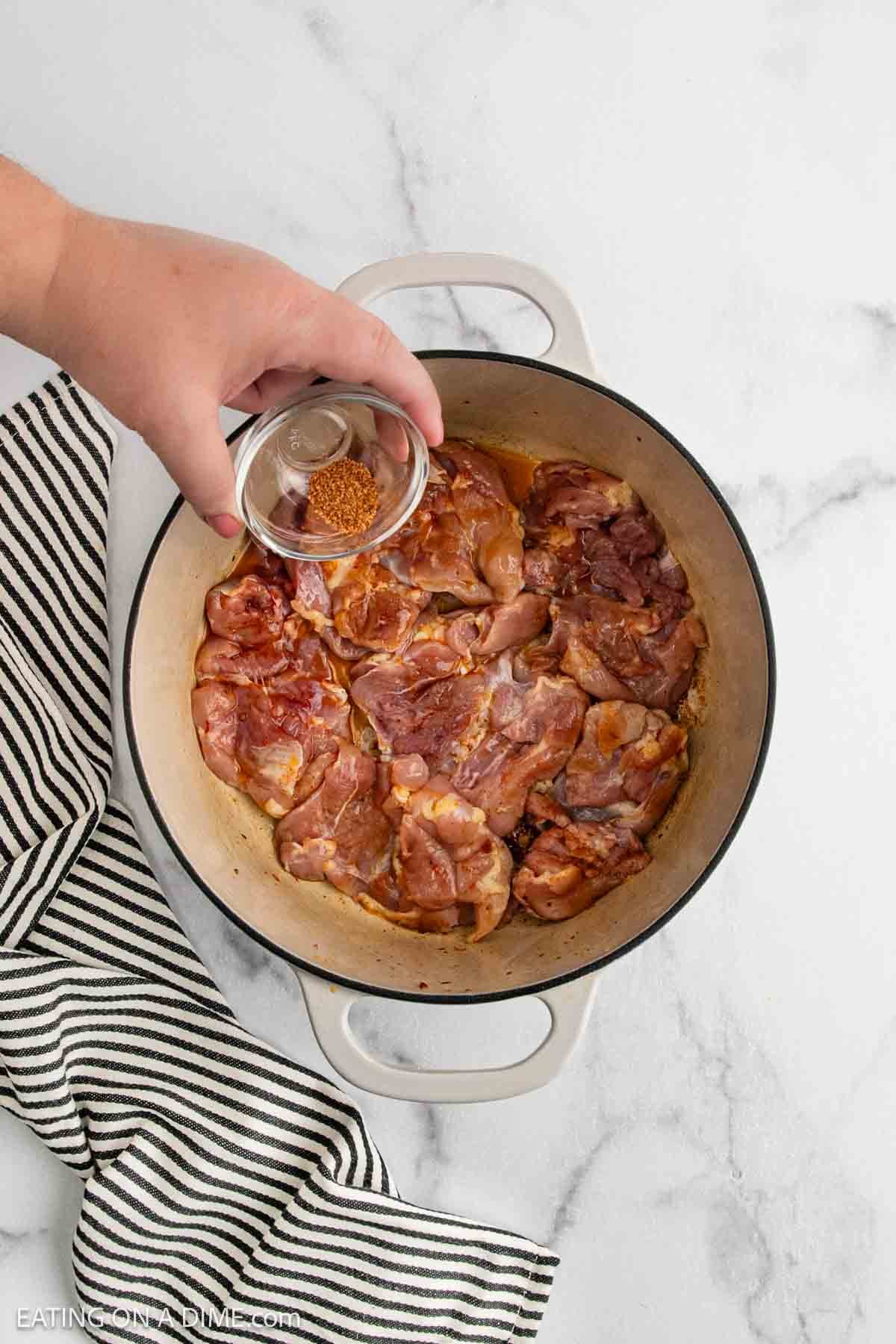 A hand sprinkles seasoning from a small glass bowl onto raw, marinated chicken pieces for a Chicken Gumbo Recipe, arranged in a large white Dutch oven on a marble surface, with a striped towel nearby.