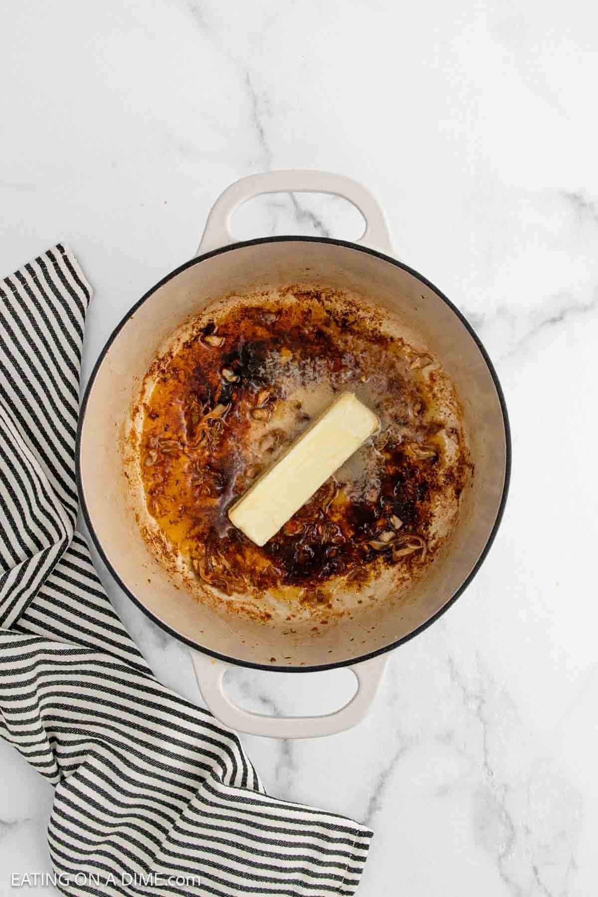 A stick of butter melting in a white Dutch oven with browned bits—perfect for starting a Chicken Gumbo Recipe—next to a black-and-white striped kitchen towel on a marble surface.