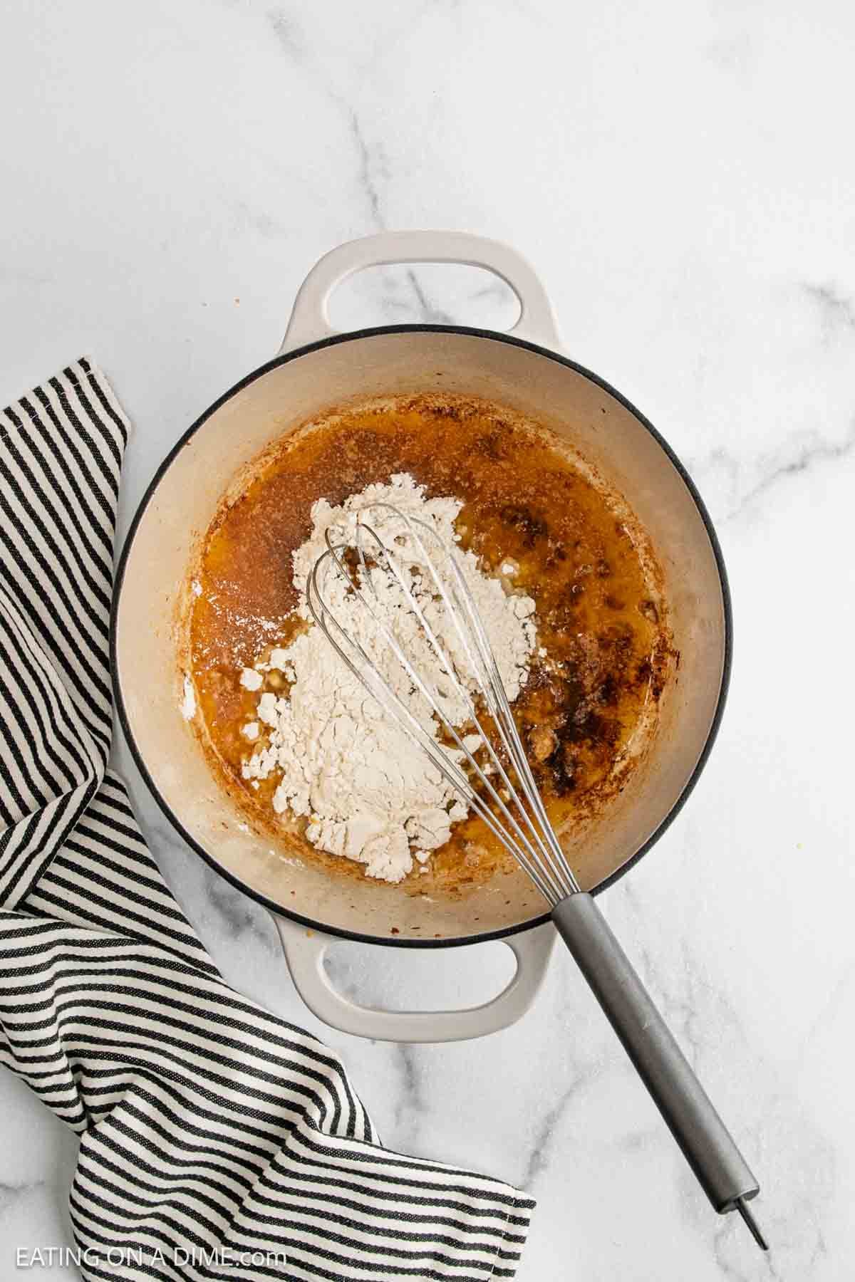 A white Dutch oven with flour being whisked into browned butter for a classic Chicken Gumbo Recipe, next to a black-and-white striped cloth on a marble countertop.