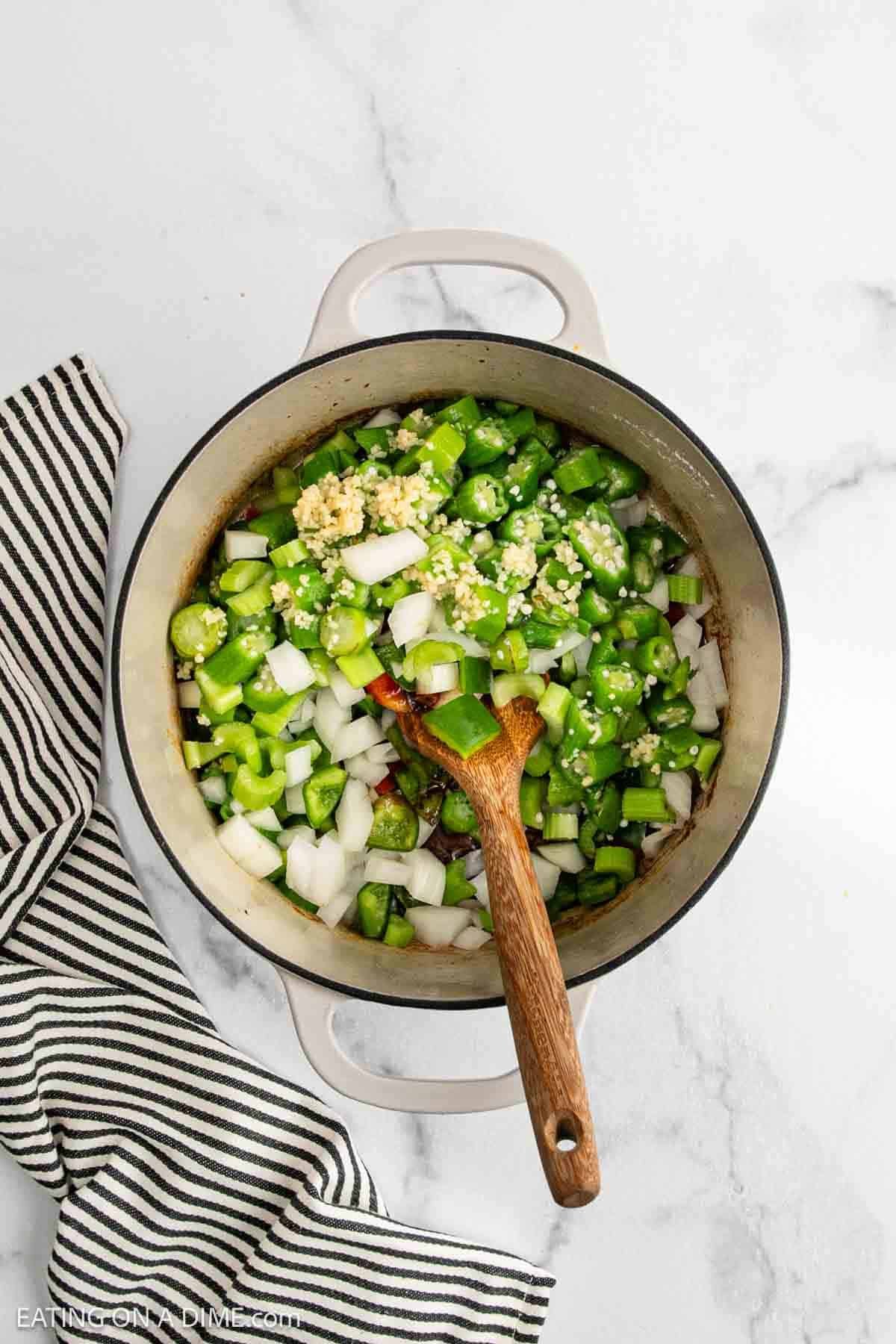 A large pot filled with chopped green bell peppers, onions, and minced garlic is being stirred for a hearty Chicken Gumbo Recipe. A black and white striped towel sits nearby on the marble countertop.