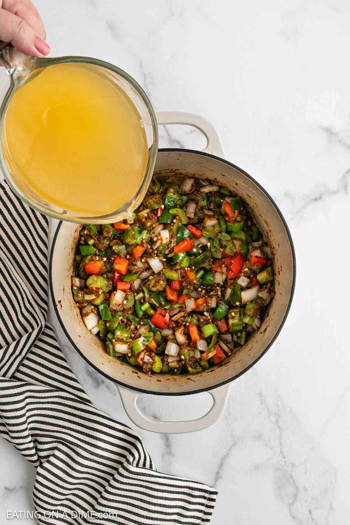 A hand pours broth from a measuring cup into a pot filled with chopped onions, peppers, and seasonings—a classic start for any Chicken Gumbo Recipe. The pot rests on a white marble surface beside a black-and-white striped towel.