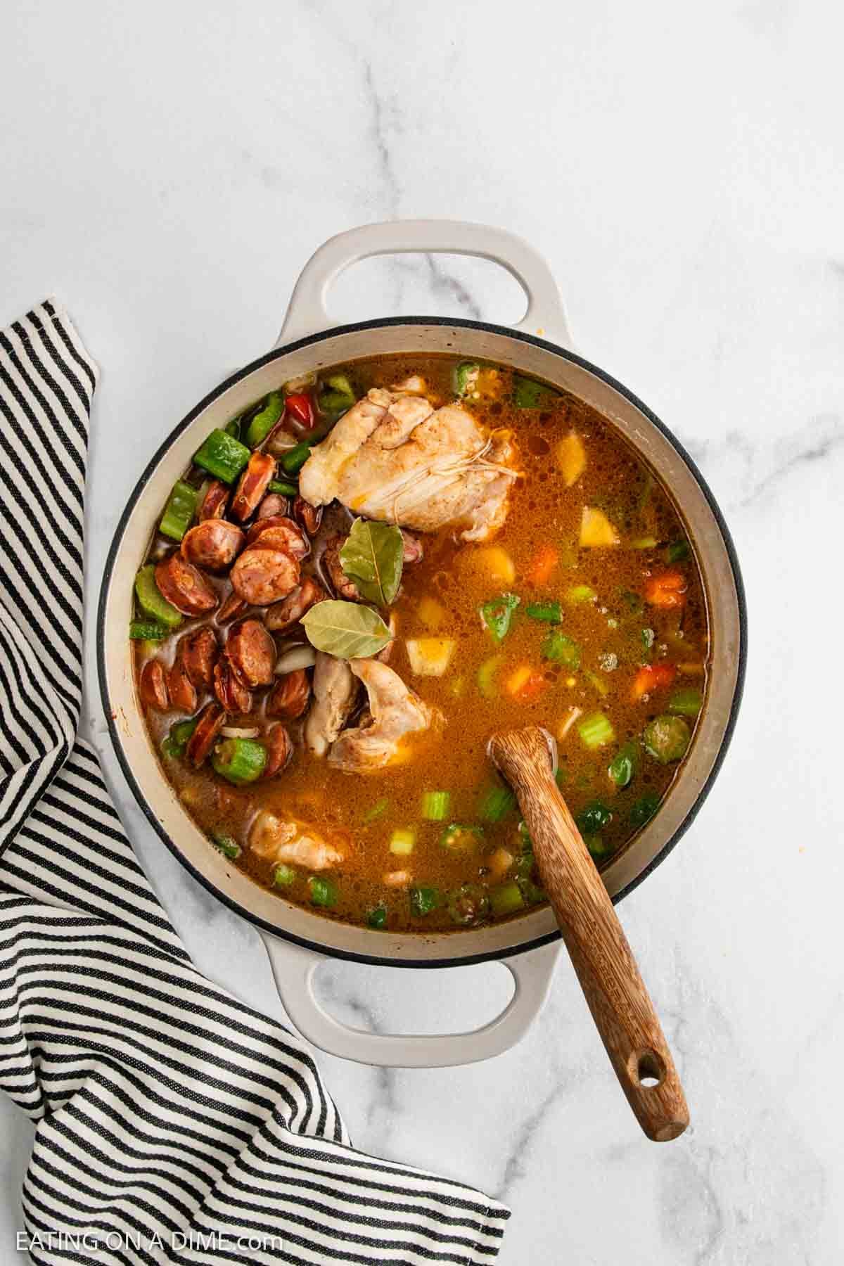 A pot of Chicken Gumbo Recipe with sausage, vegetables, and bay leaves sits on a marble surface next to a black and white striped towel. A wooden spoon rests inside the pot.