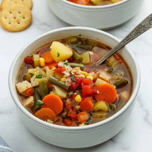 A bowl of Chicken Vegetable Soup with carrots, potatoes, corn, green beans, celery, and diced tomatoes is served with a spoon. Crackers are placed beside the bowl on a white surface.