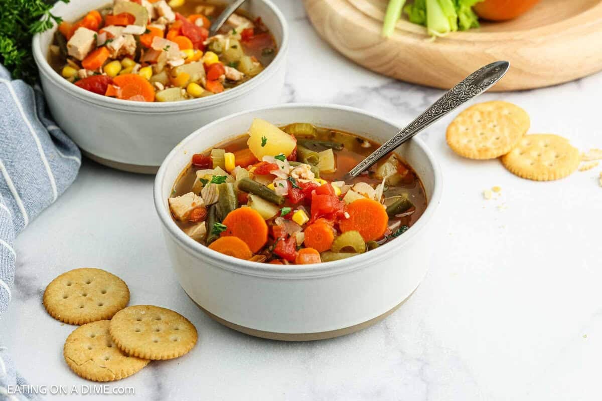Two bowls of hearty vegetable soup with carrots, potatoes, corn, and green beans sit ready with spoons. Round butter crackers are scattered nearby. In the background, a wooden board holds celery and an orange.