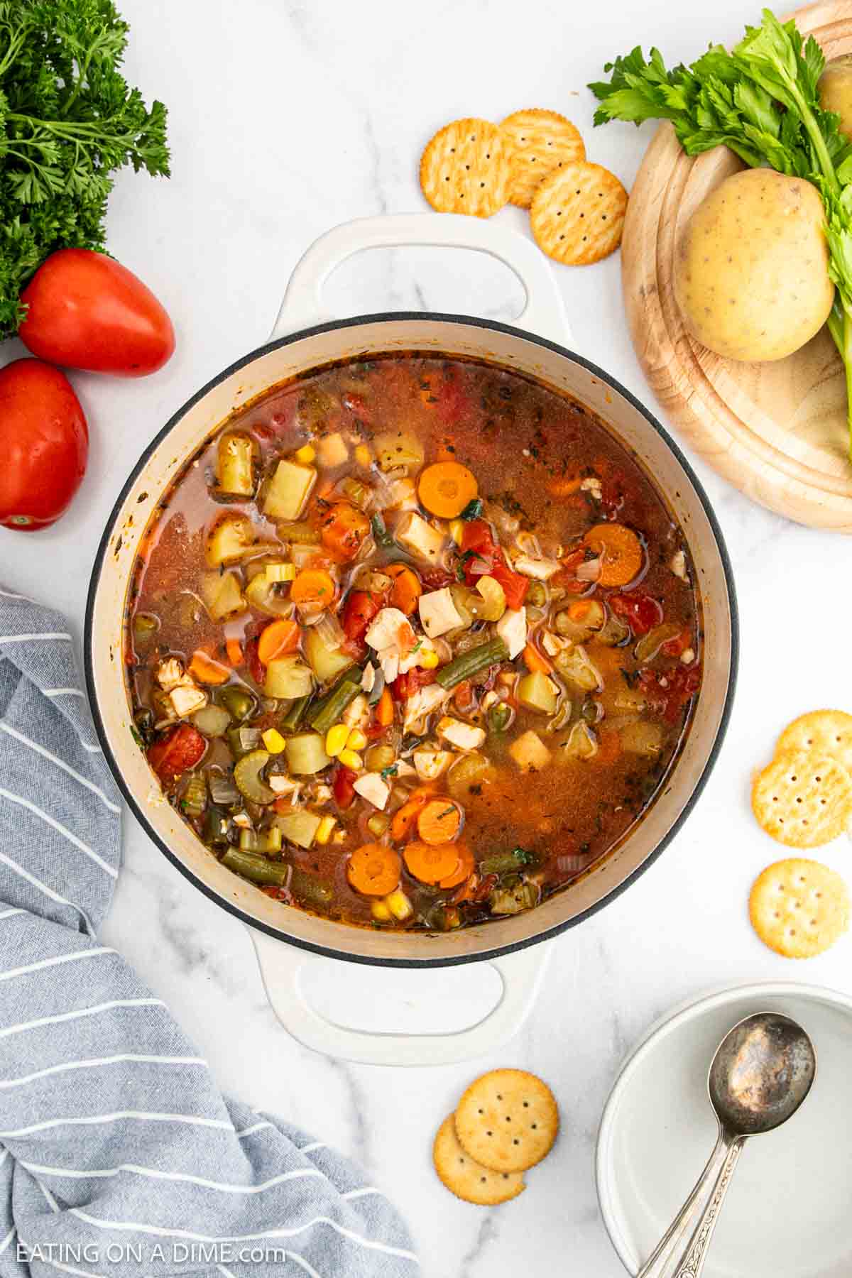 A pot of chicken vegetable soup with carrots, celery, corn, tomatoes, and pasta sits on a white countertop, surrounded by crackers, fresh vegetables, herbs, a spoon, and a blue-striped towel.