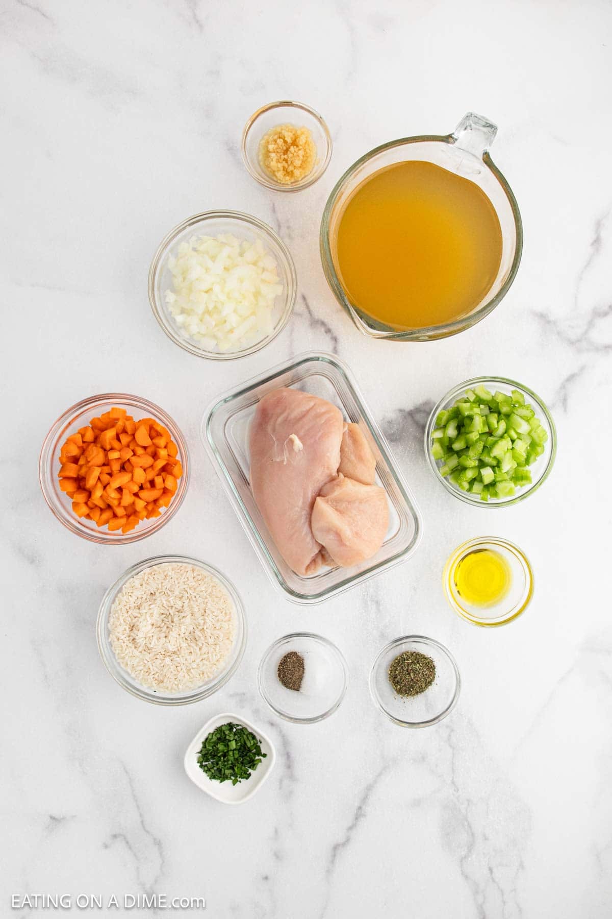 Top-down view of ingredients for chicken and rice soup, featuring raw chicken breasts, chopped onions, sliced carrots, diced celery, rice, minced garlic, chicken broth, olive oil, salt, pepper, and fresh herbs on a white surface.