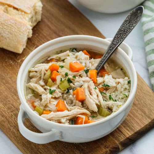 A comforting bowl of Chicken and Rice Soup with carrots and celery, garnished with herbs, sits on a wooden board beside slices of bread. A spoon rests in the bowl, with a striped towel nearby for a cozy touch.