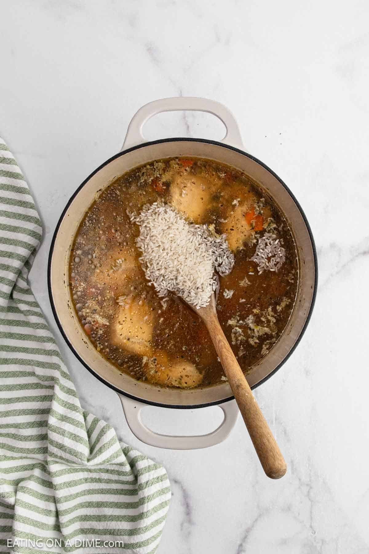 A white pot filled with Chicken and Rice Soup, featuring tender chicken pieces and uncooked rice being stirred with a wooden spoon, sits on a white marble surface next to a green striped towel.