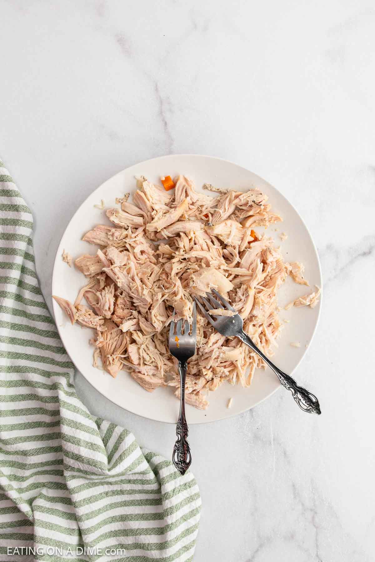 A white plate with shredded chicken and two forks on a marble surface, next to a green and white striped towelโperfect for adding to chicken soup or rice soup recipes.