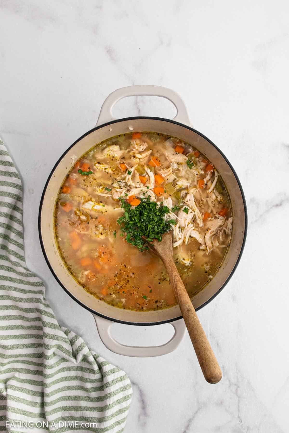 A pot of homemade Chicken and Rice Soup with shredded chicken, carrots, rice, and herbs is being stirred with a wooden spoon. Fresh chopped parsley tops the soup, and a striped kitchen towel sits beside it on the marble countertop.