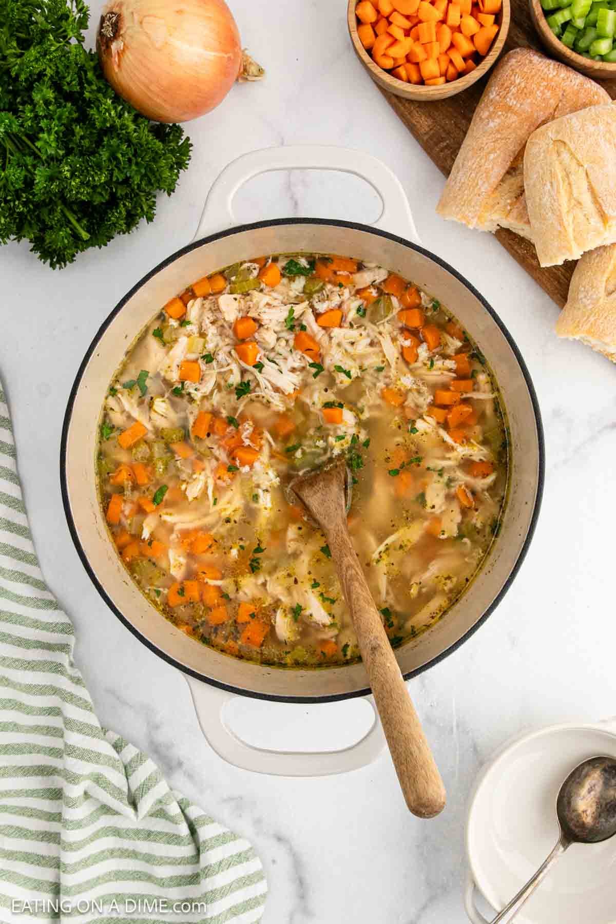 A pot of homemade Chicken and Rice Soup with shredded chicken, carrots, rice, and herbs sits on a marble countertop with a wooden spoon. Surrounding the pot are bread, chopped vegetables, parsley, and an onion.