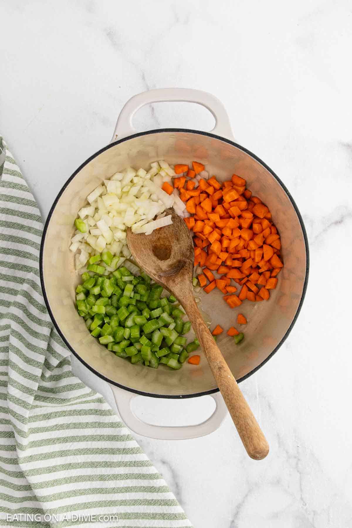 A white pot holds chopped onions, carrots, and celeryโclassic ingredients for Chicken and Rice Soupโdivided into sections with a wooden spoon on top. A green and white striped towel rests nearby on the marble surface.