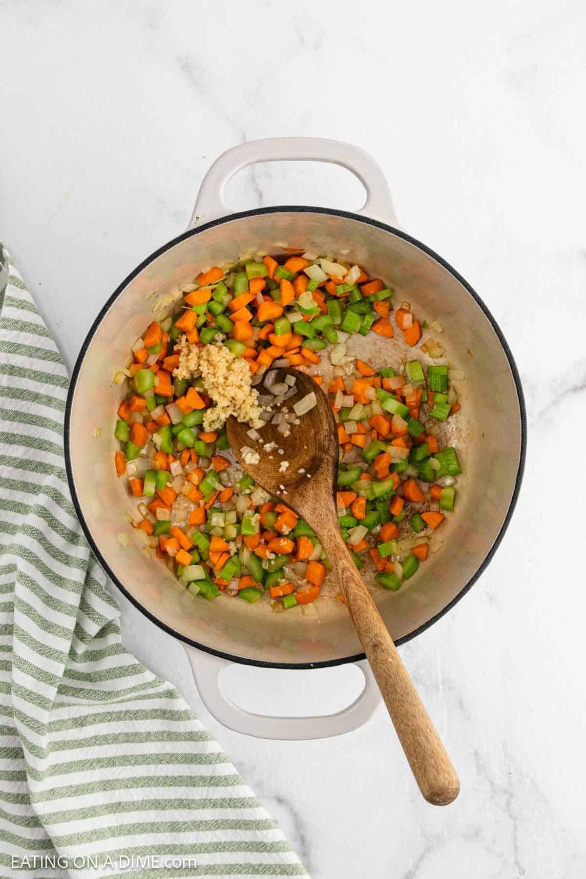 A pot with chopped carrots, celery, onions, and minced garlic being sautรฉed for Chicken and Rice Soup, stirred with a wooden spoon. A green and white striped towel rests beside the pot on a white countertop.