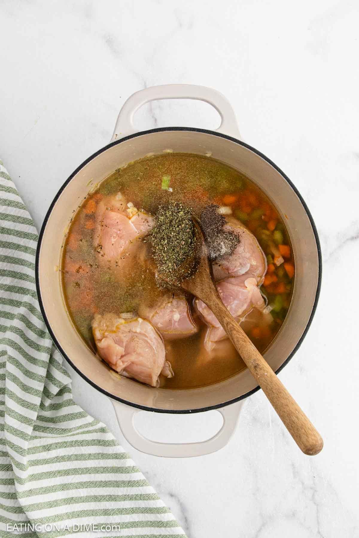 A pot filled with raw chicken, broth, chopped carrots, celery, and seasonings for Chicken and Rice Soup is being stirred with a wooden spoon on a white surface beside a green and white striped towel.