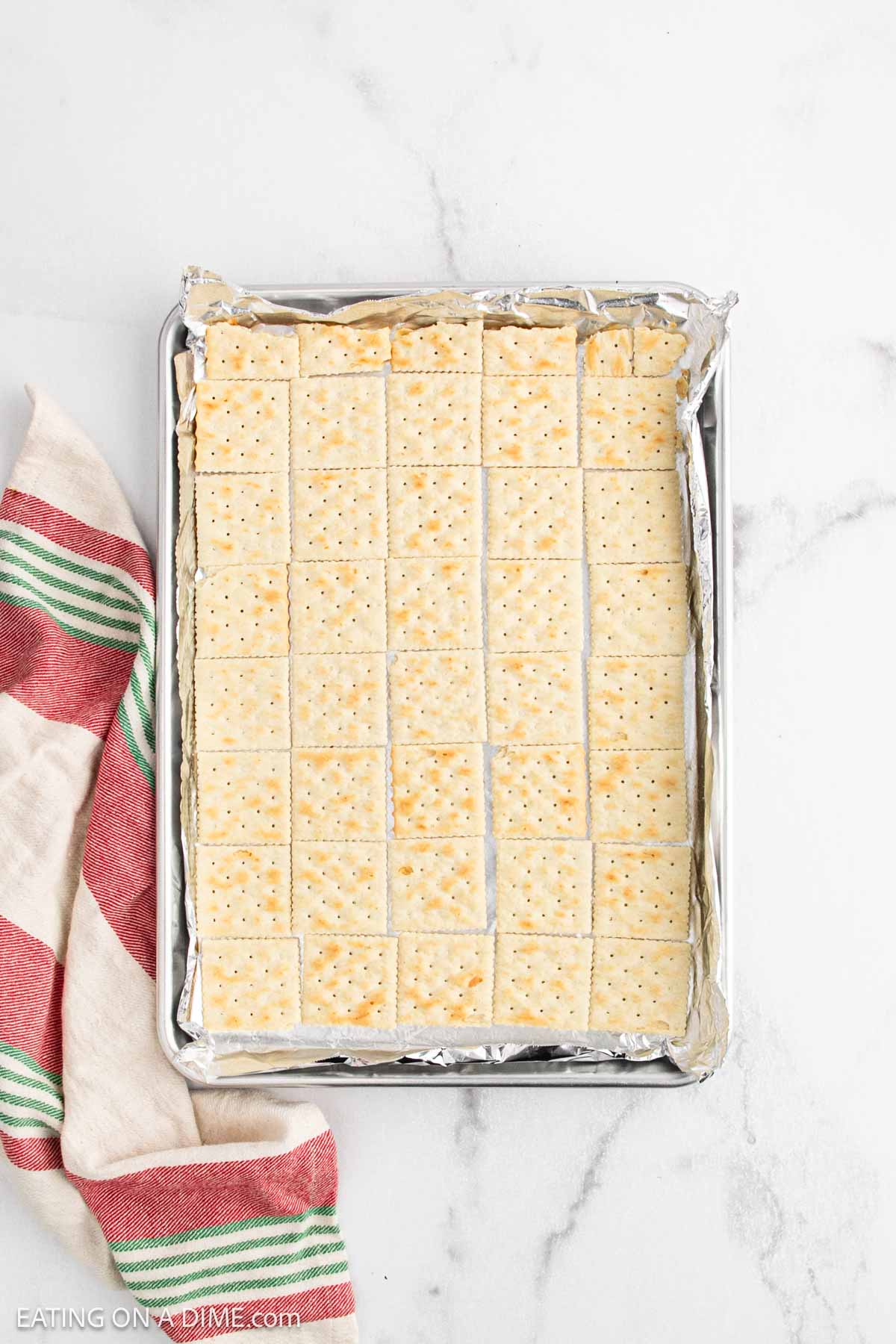 A baking sheet lined with foil holds a single layer of saltine crackers, ready to become delicious Christmas Crack. A red, green, and cream striped kitchen towel sits beside the tray on a white marble surface.