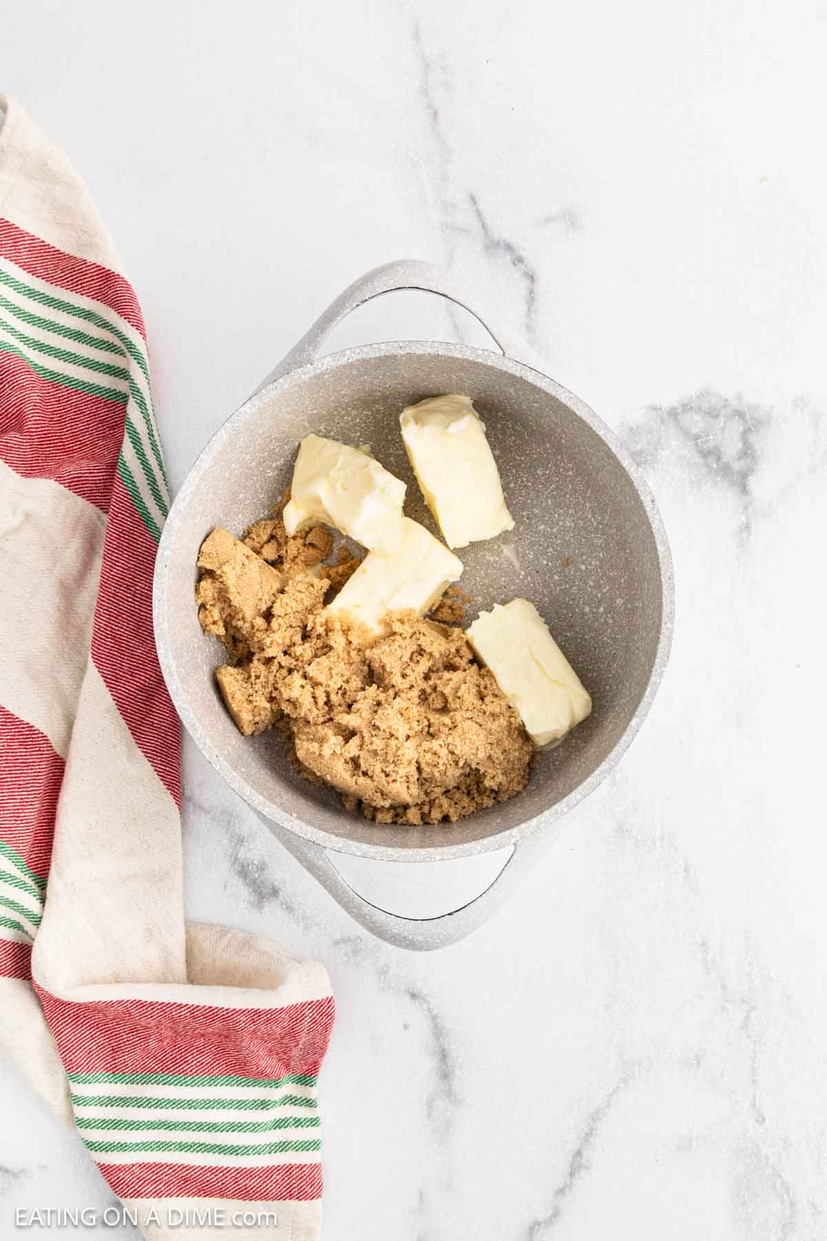 A metal bowl on a marble countertop holds chunks of butter and packed brown sugar—perfect for starting Christmas Crack. A red, green, and cream striped kitchen towel lies beside the bowl.