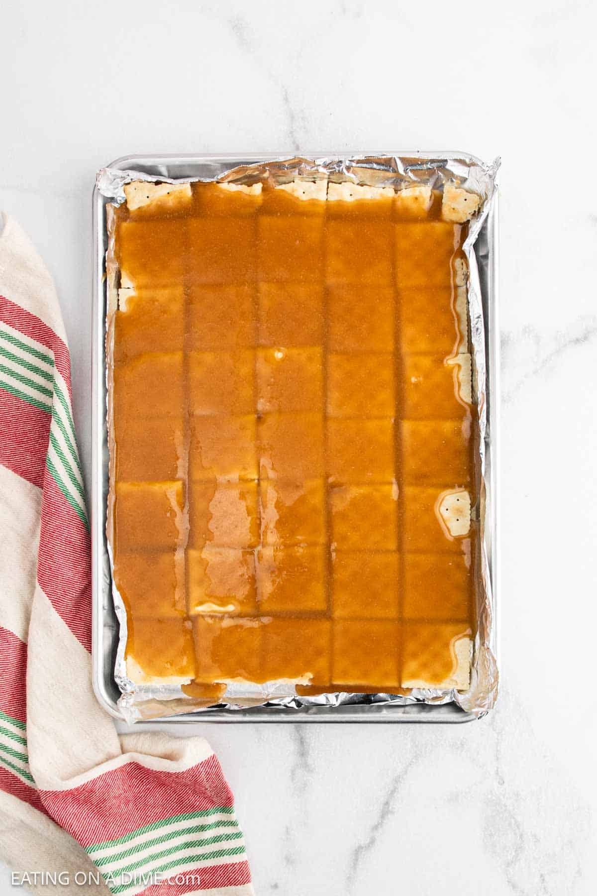 A baking sheet lined with foil holds a batch of Christmas Crack caramel squares, partially cut into a grid. A red and green striped kitchen towel lies beside the pan on a white marble surface.