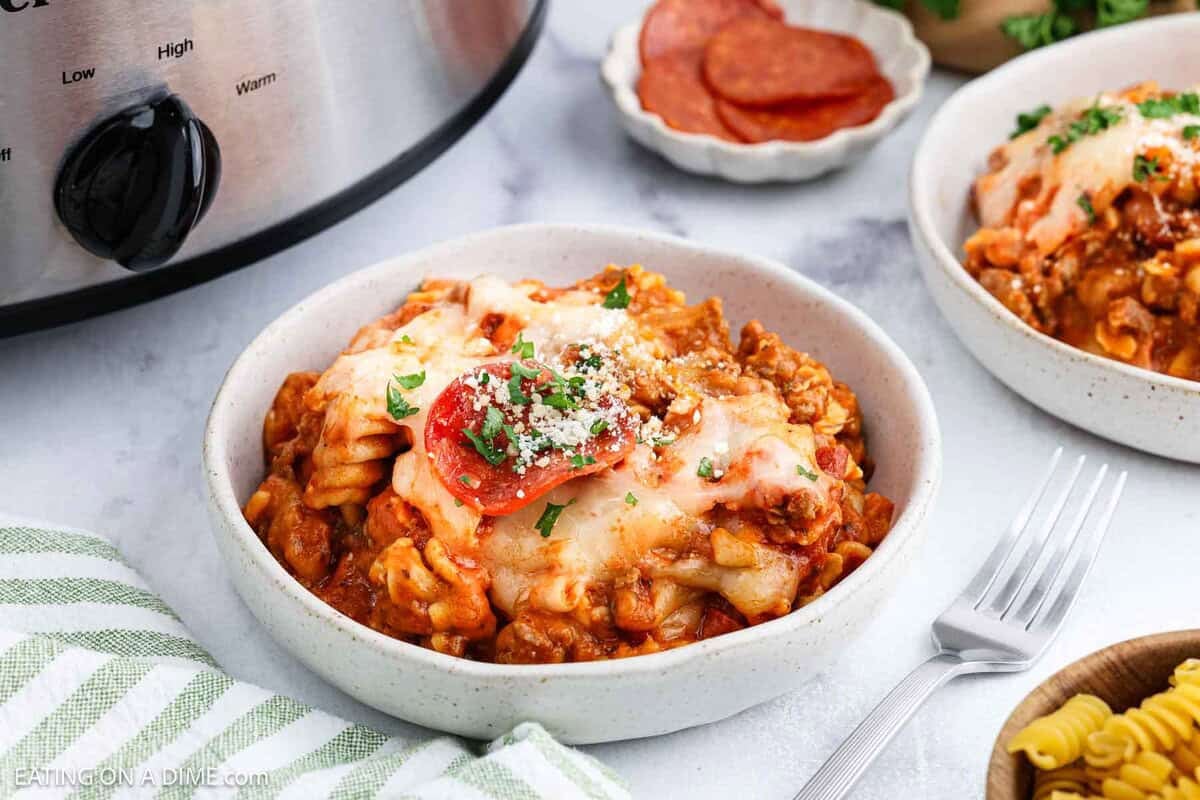 A bowl of Pizza Casserole topped with melted cheese, pepperoni, and parsley sits on a table beside a fork. In the background are another bowl, a Crock Pot, and a small plate of pepperoni.
