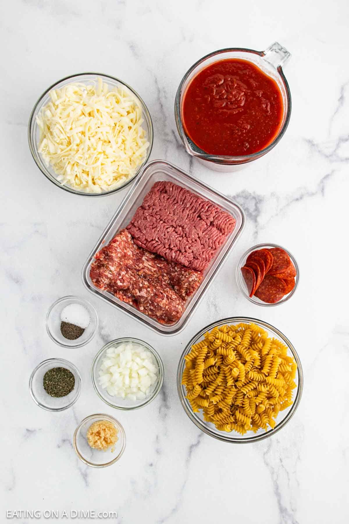 Top-down view of Crock Pot Pizza ingredients for a pizza casserole—shredded cheese, tomato sauce, ground beef and sausage, pepperoni slices, rotini pasta, onions, garlic, dried herbs, salt, and pepper.