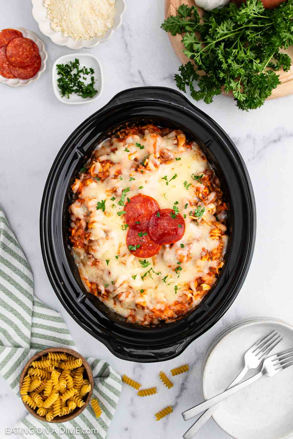 A Crock Pot filled with cheesy baked pasta, topped with pepperoni slices and parsley, transforms into a delicious Pizza Casserole. Surrounded by plates, dried pasta, grated cheese, fresh parsley, and a bowl of pepperoni on a marble surface.