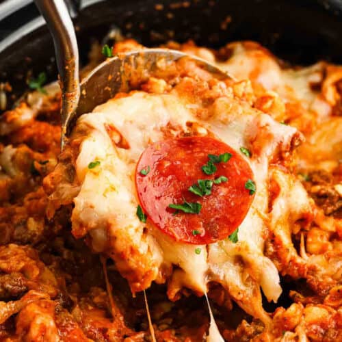 A serving spoon lifts a cheesy, baked Crock Pot Pizza Casserole with melted mozzarella, ground meat, tomato sauce, and a slice of pepperoni, topped with chopped parsley.