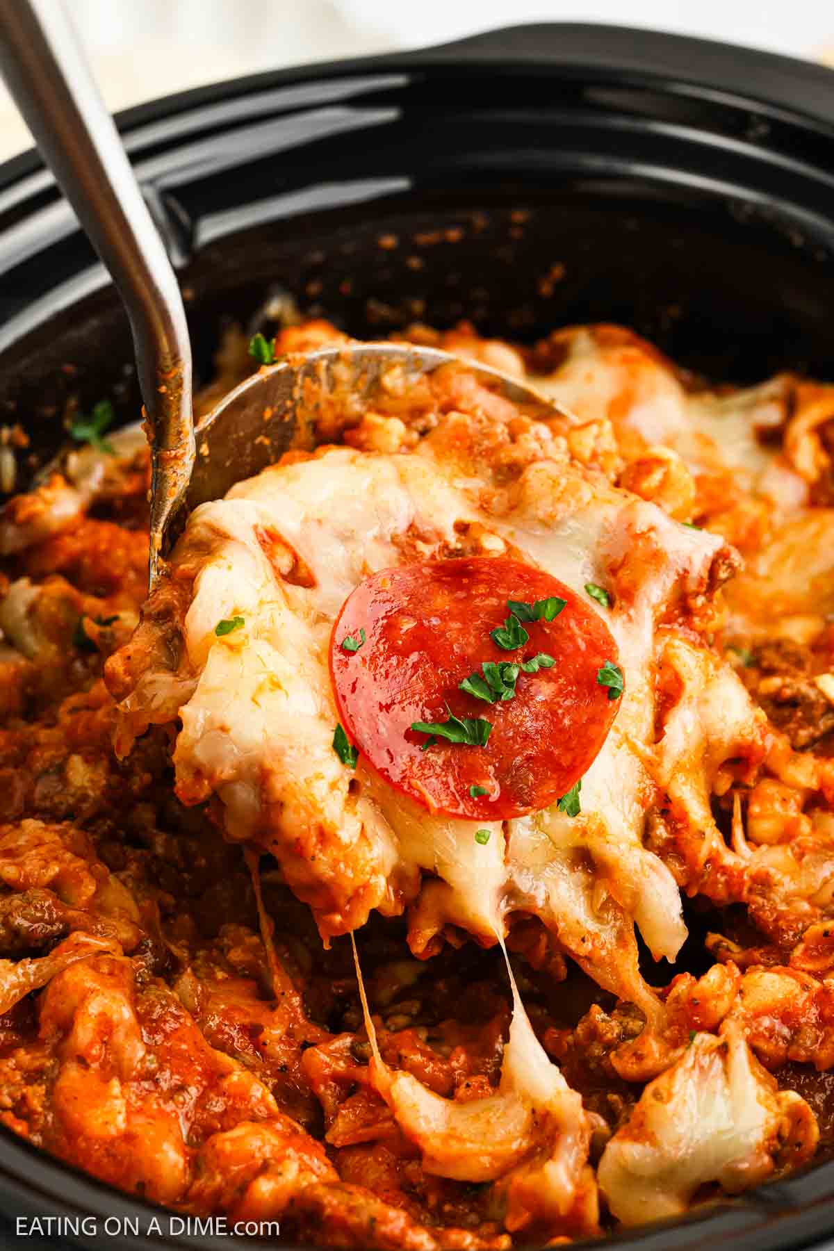 A close-up of a cheesy Pizza Casserole being scooped from a Crock Pot, featuring melted cheese, tomato sauce, noodles, and pepperoni on top, all garnished with chopped herbs.
