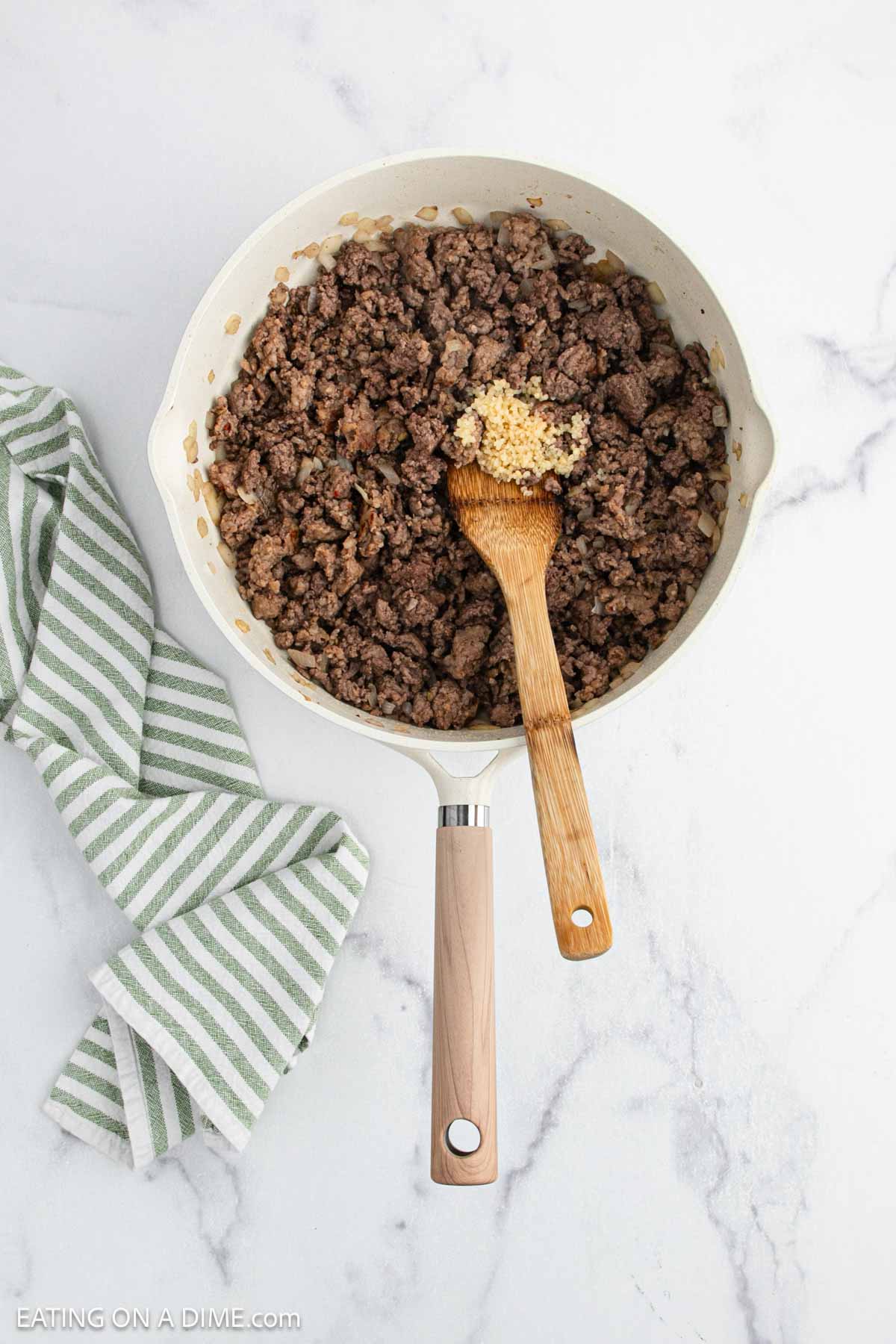 Cooked ground beef with chopped onions and minced garlic in a white skillet, stirred with a wooden spoon—perfect as a savory base for Crock Pot Pizza Casserole—next to a green and white striped towel on a marble countertop.