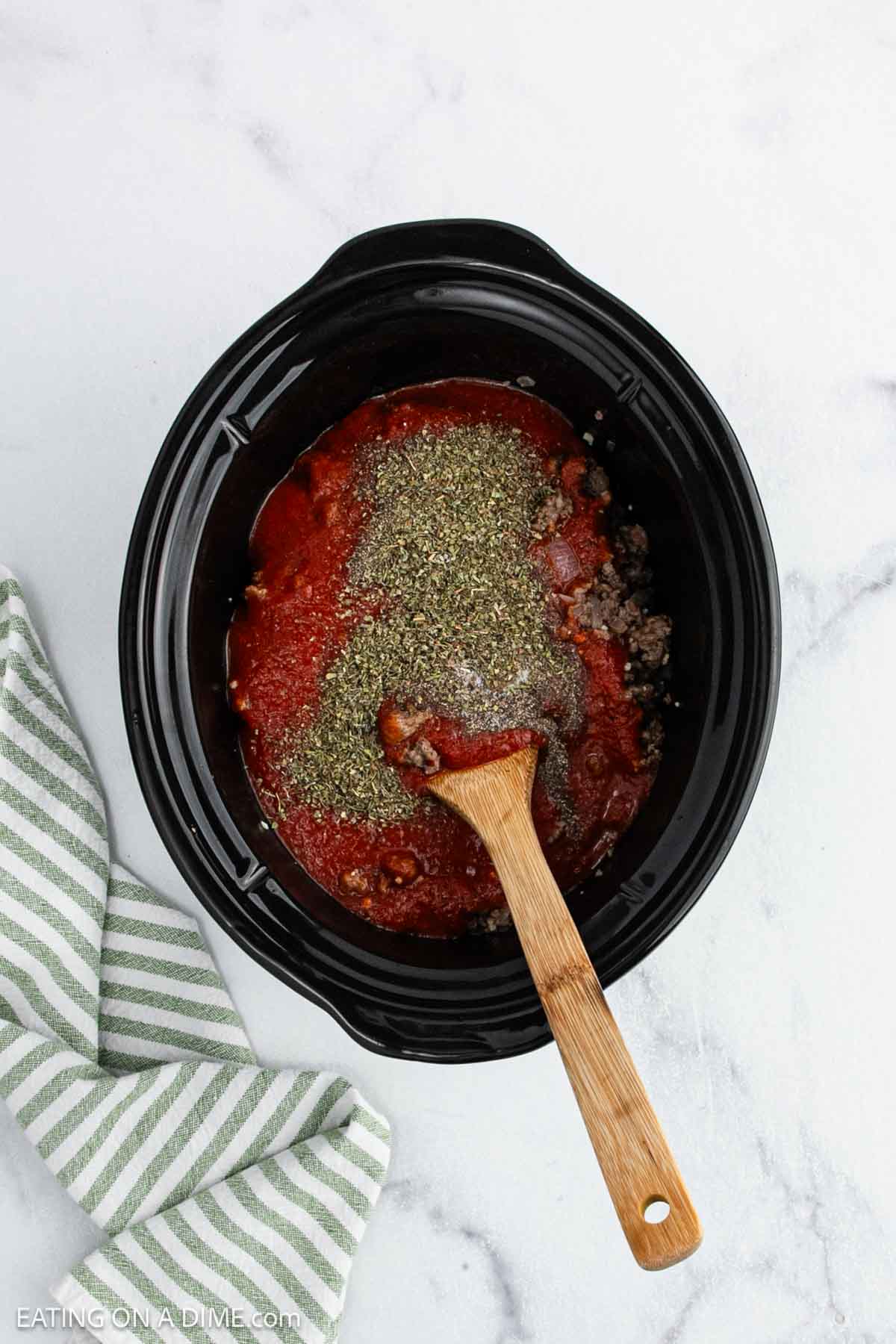 A Crock Pot filled with ground beef, tomato sauce, and dried herbs is being stirred with a wooden spoon—perfect for making a hearty Pizza Casserole. A green and white striped cloth rests beside it on the white marble surface.