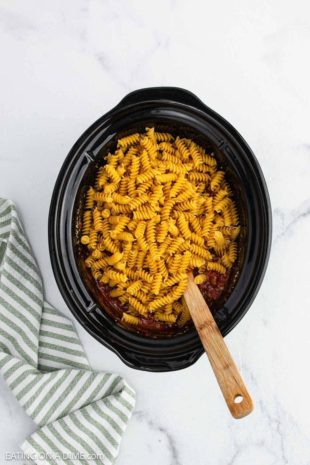 A black Crock Pot filled with dry rotini pasta and a wooden spoon resting inside. A green and white striped kitchen towel is placed nearby on a white marble surface, ready for your next Pizza Casserole creation.