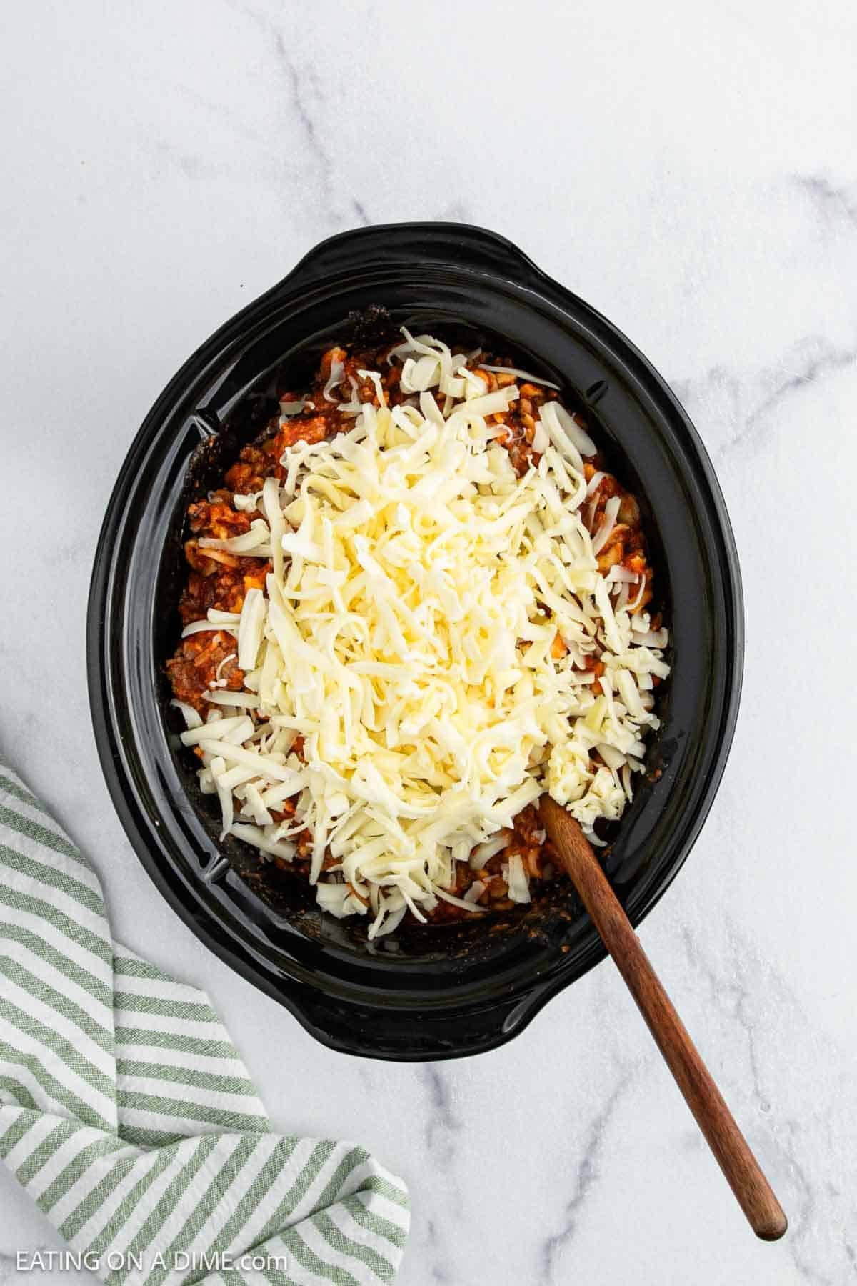 Overhead view of a Crock Pot filled with a cheesy pasta dish topped with shredded cheese, with a wooden spoon inside. A green and white striped towel rests nearby on a marble surface—perfect for making Crock Pot Pizza Casserole.