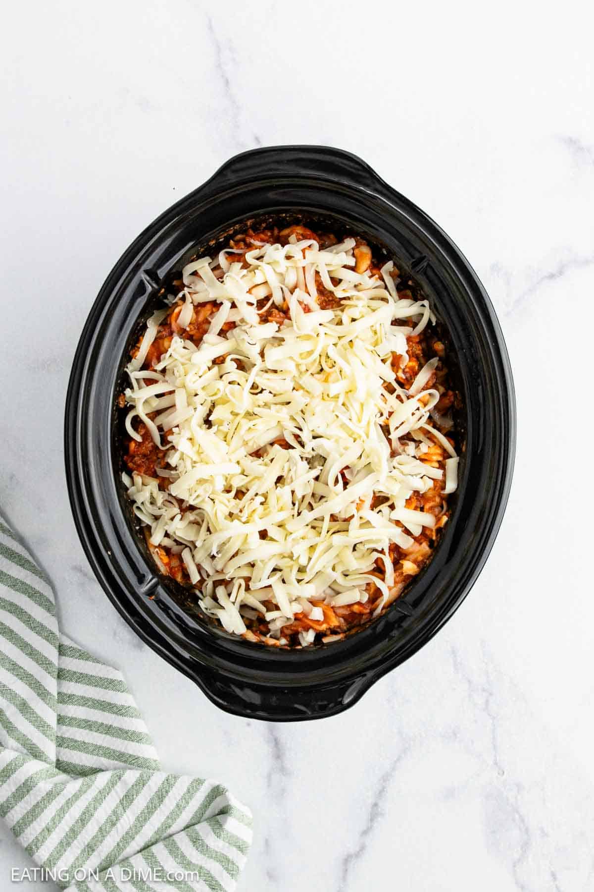 A black Crock Pot filled with a layered pizza casserole topped with shredded cheese sits on a white marble surface next to a green striped cloth.