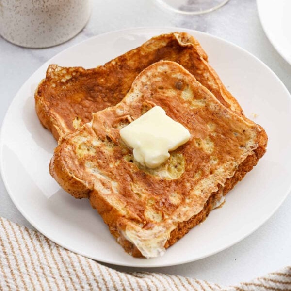 Two slices of Eggnog French Toast topped with a pat of melting butter, served on a white plate with a beige-striped napkin nearby.