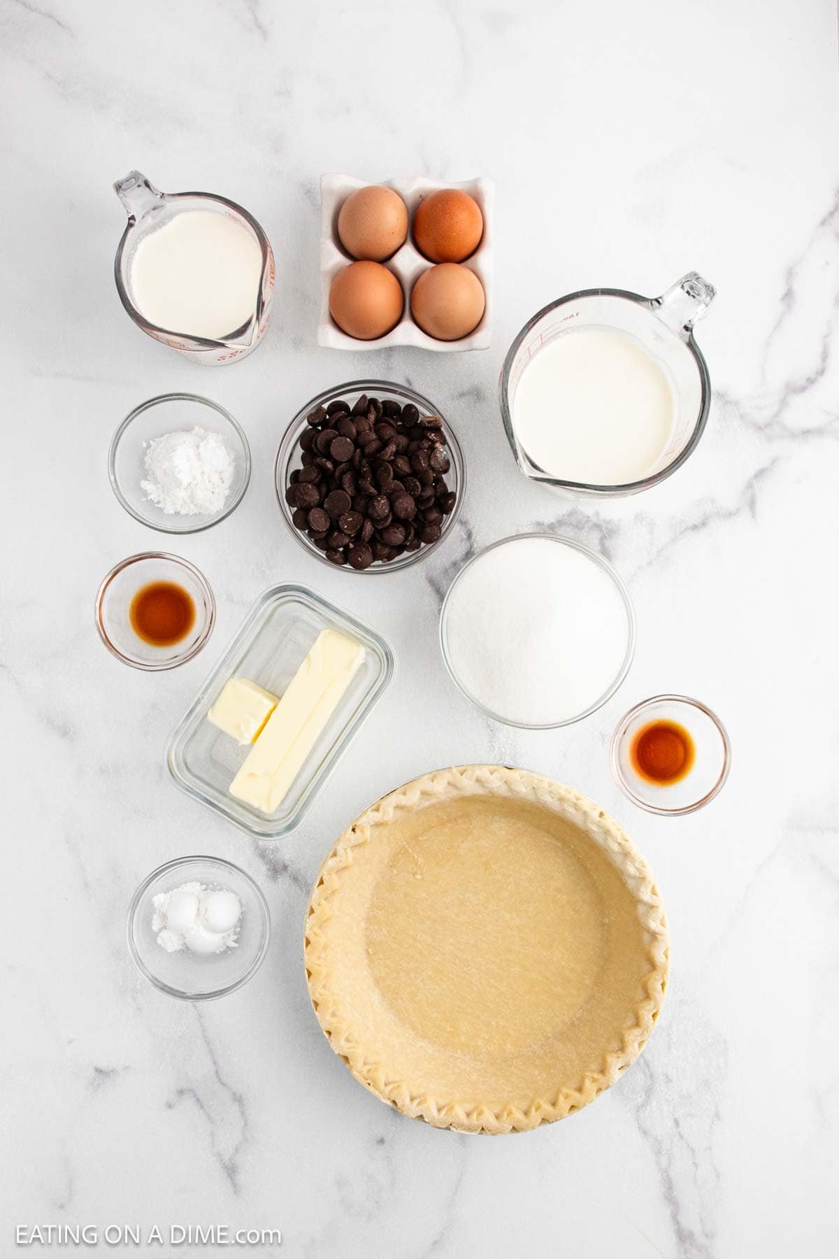 Overhead view of ingredients for a French Silk Pie: eggs, chocolate chips, sugar, butter, milk, cream, vanilla extract, cornstarch, salt, a pie crust, and baking powder arranged on a white marble surface.