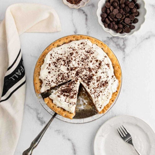 A French Silk Pie topped with chocolate shavings sits on a marble surface. One slice is missing, and a pie server rests beside the empty space. Nearby are a white plate, a fork, a bowl of chocolate chips, and a folded napkin.
