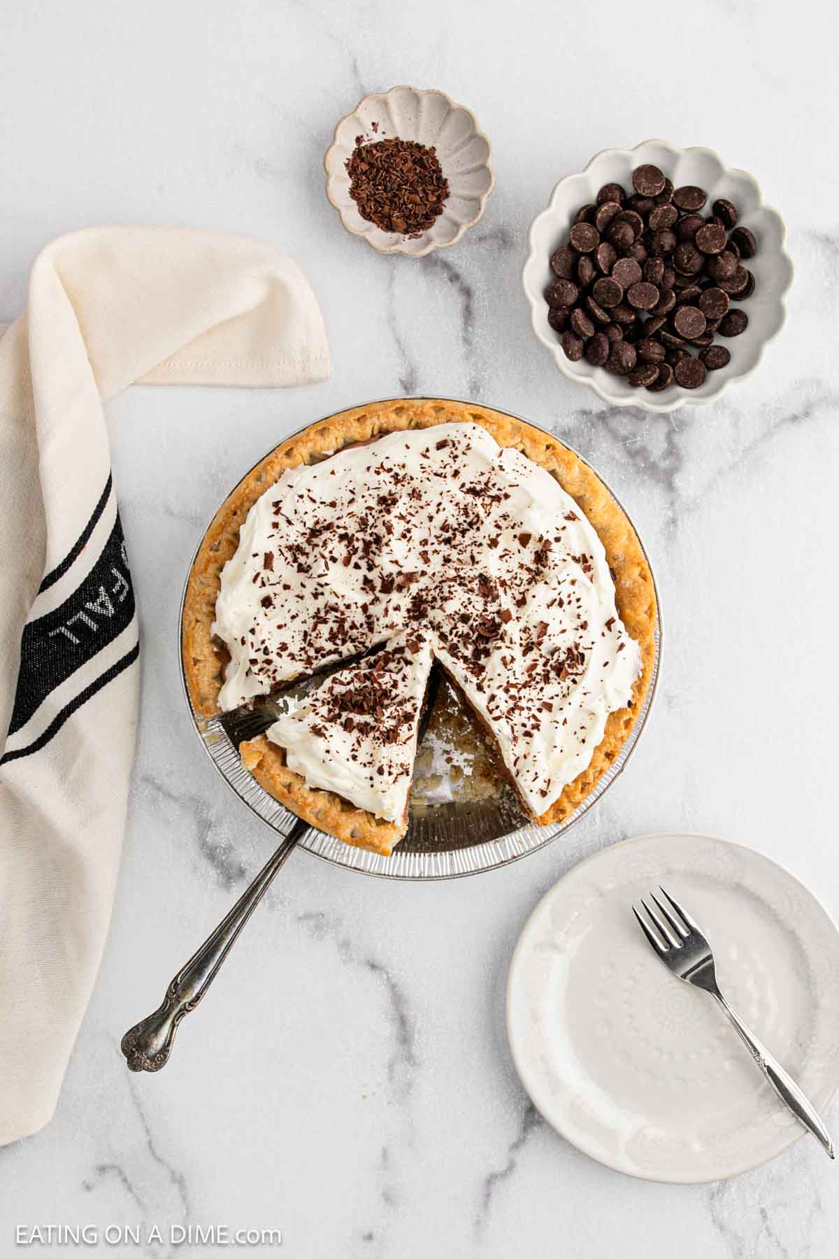 A decadent French Silk Pie topped with whipped cream and chocolate shavings sits on a marble surface. A slice is cut out on a plate with a fork, surrounded by bowls of chocolate chips and extra chocolate shavings.