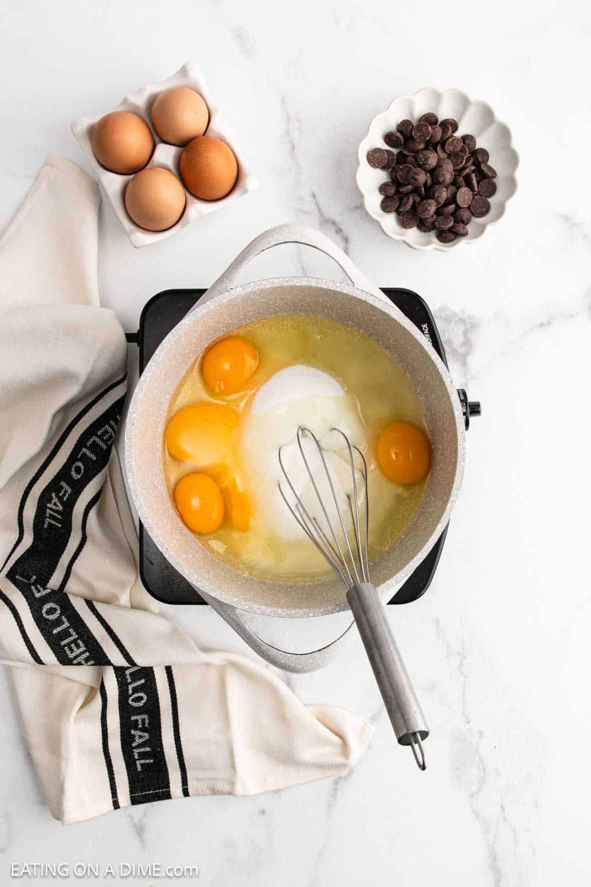 A pot with eggs, sugar, and a whisk sits on a stovetop—steps to making French Silk Pie. Nearby are chocolate chips, more eggs, and a cream-colored towel with black stripes on a white marble surface.