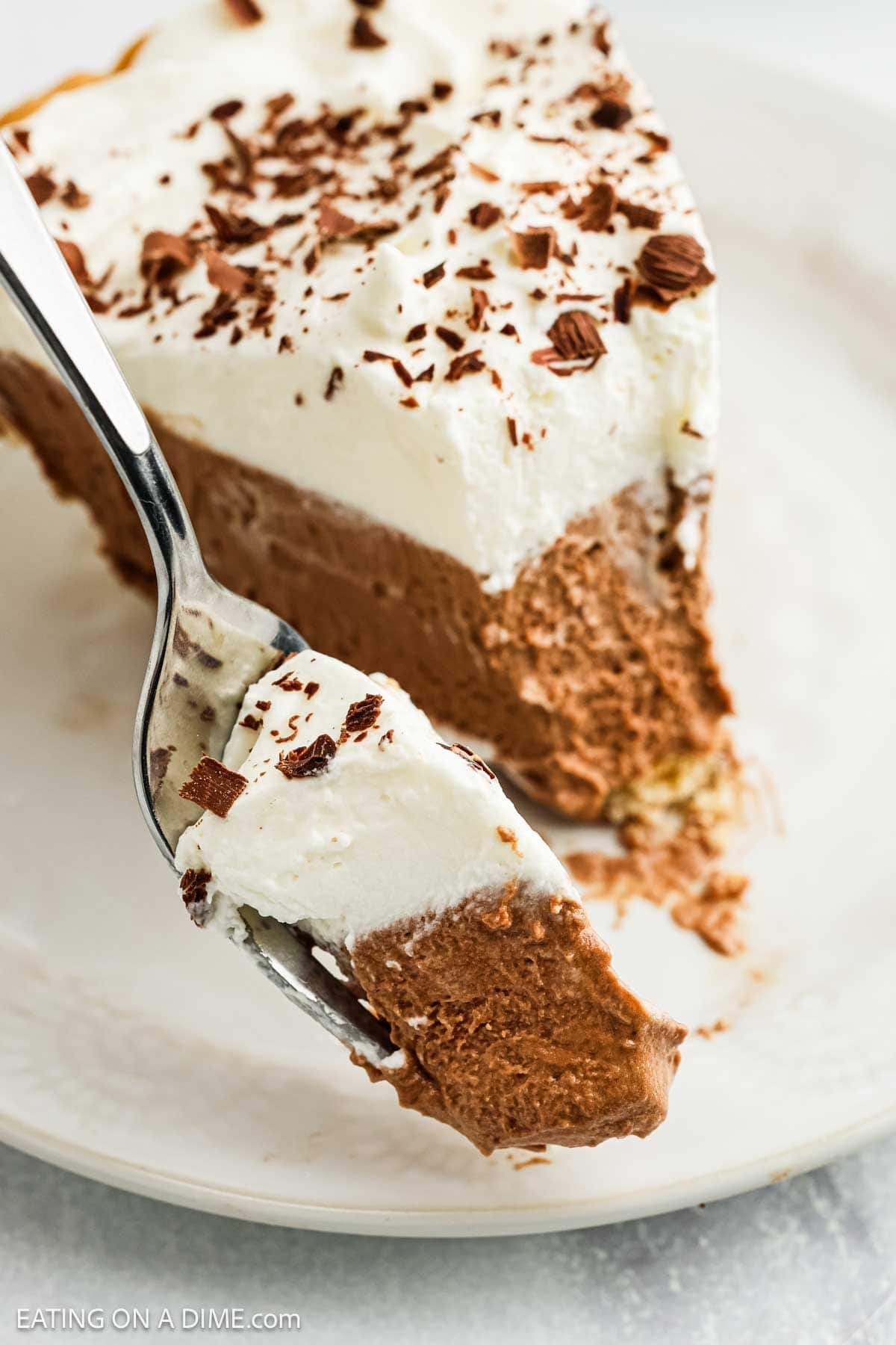 A close-up of a fork holding a bite of French Silk Pie topped with whipped cream and chocolate shavings, with the rest of the luscious pie slice on a white plate in the background.