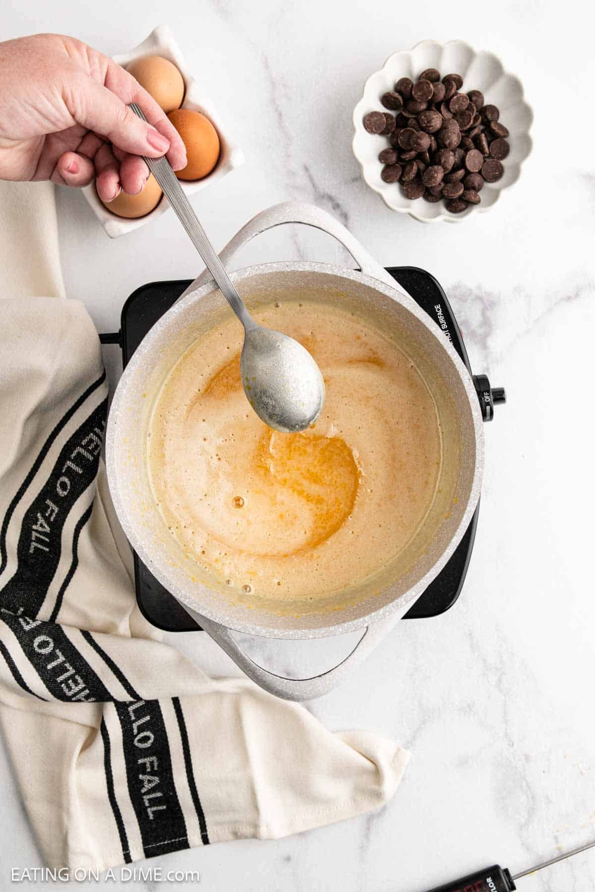 A hand stirs a creamy mixture for French Silk Pie in a saucepan on the stovetop. Nearby are a dish with eggs, a bowl of chocolate pieces, a "HELLO FALL" kitchen towel, and a thermometer.