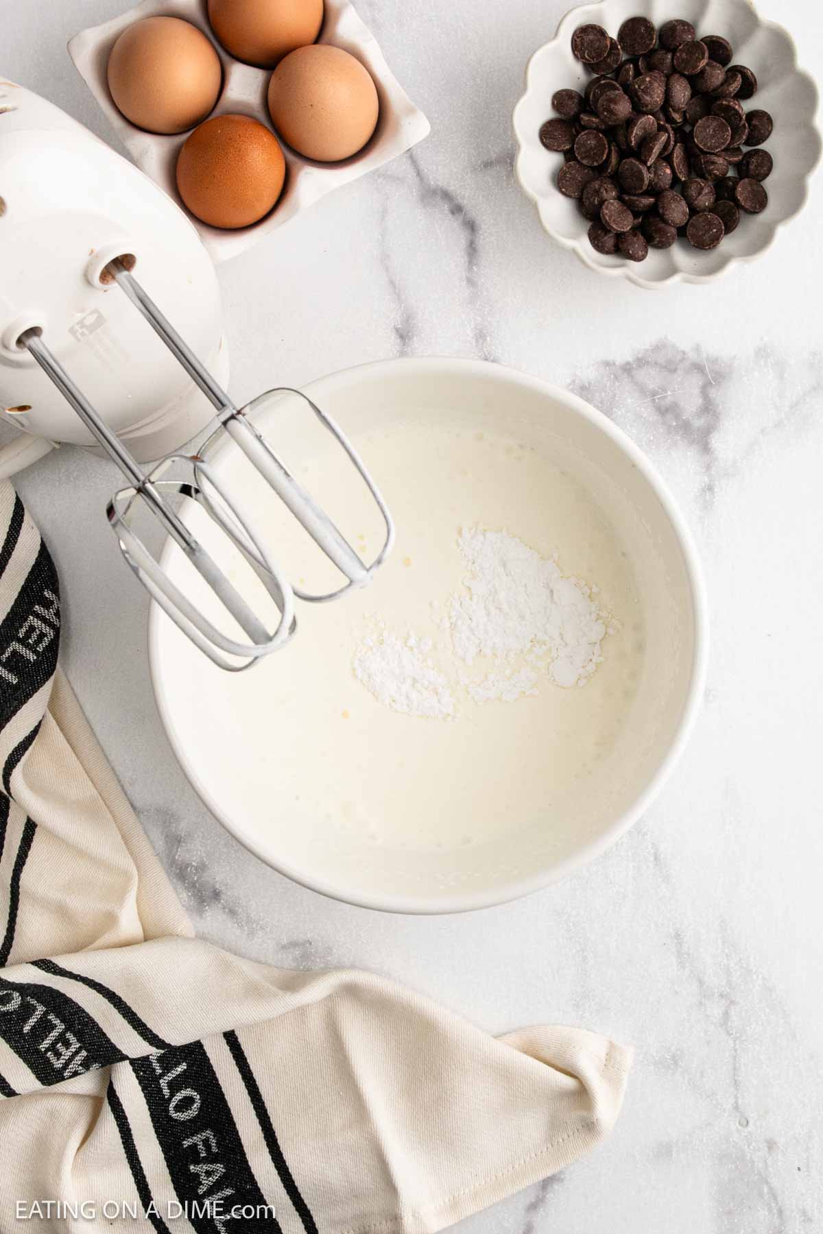 A mixing bowl with cream and powdered sugar for French Silk Pie sits next to a mixer, eggs in a dish, chocolate chips in a bowl, and a striped kitchen towel on a marble countertop.