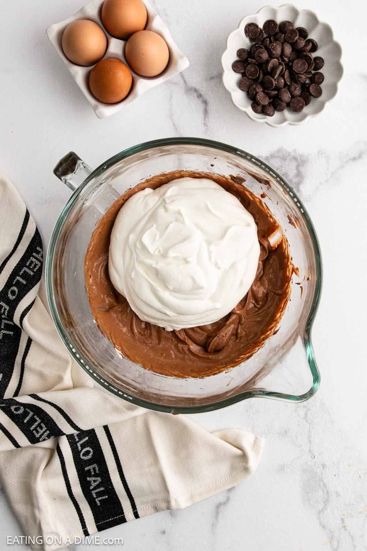 A glass mixing bowl with chocolate mixture and whipped cream, perfect for French Silk Pie, sits on a marble counter, surrounded by a dish of eggs, a bowl of chocolate chips, and a cream-colored towel with black stripes.