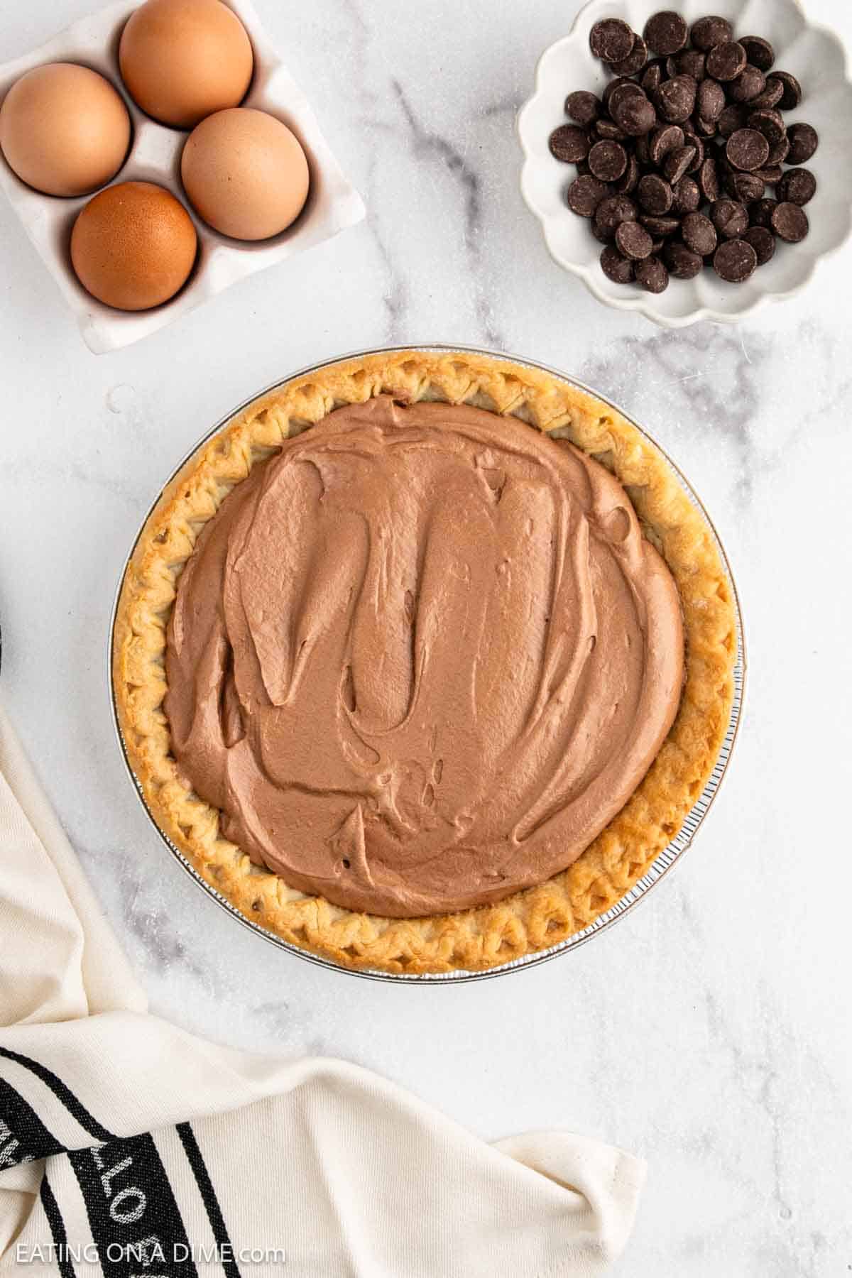 A French Silk Pie with a flaky crust sits on a marble surface. Nearby are a dish of eggs, a bowl of chocolate chips, and a white cloth with a black stripe.