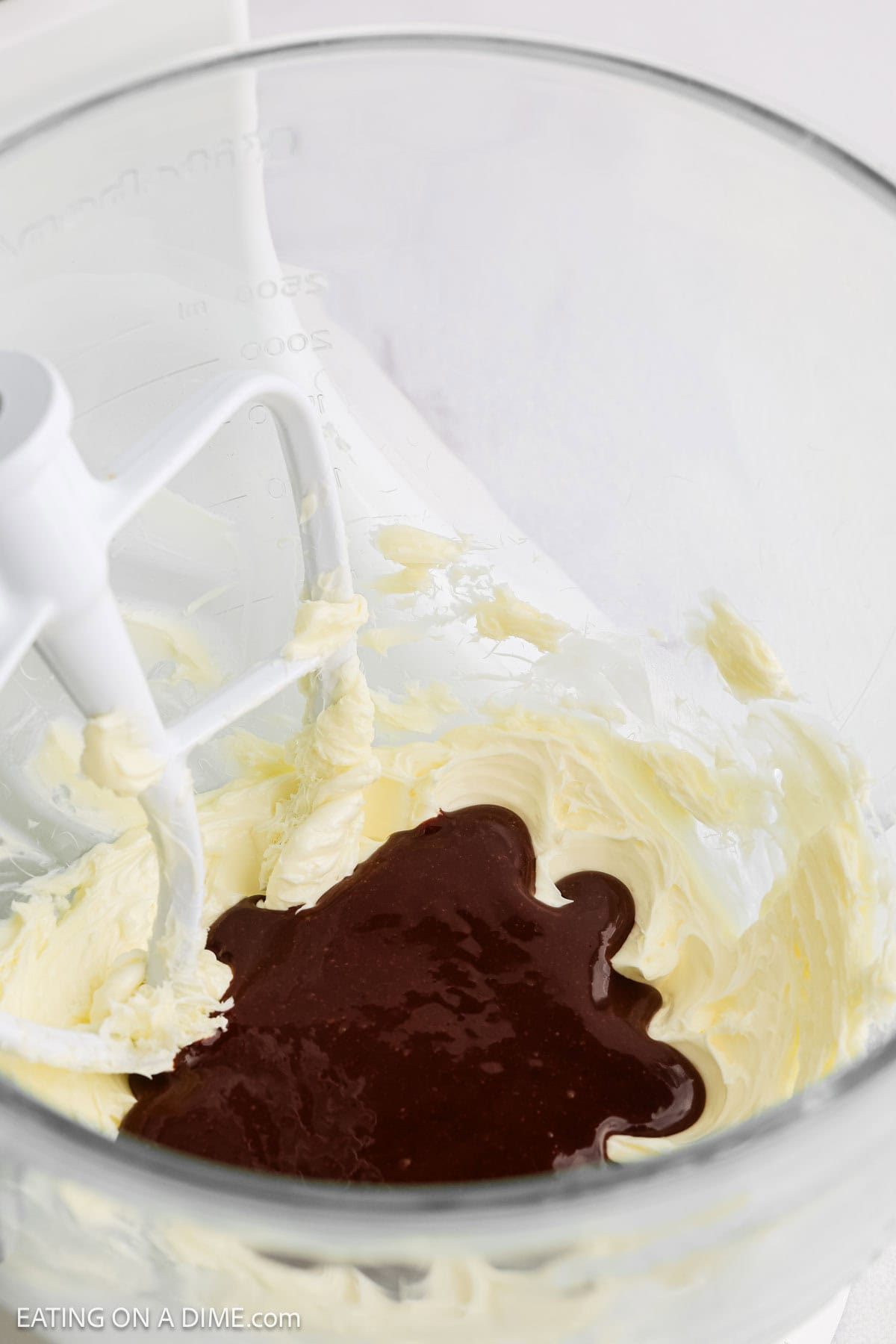 A glass mixing bowl with softened butter and dark brown liquid, possibly melted chocolate for French Silk Pie, being mixed with a white paddle attachment of a stand mixer.