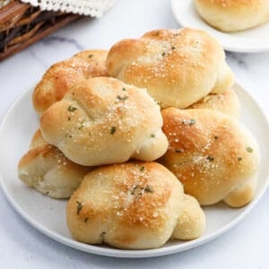 A white plate piled with golden-brown Garlic Knots sprinkled with herbs and grated cheese, set on a light surface with another delicious garlic knot in the background.