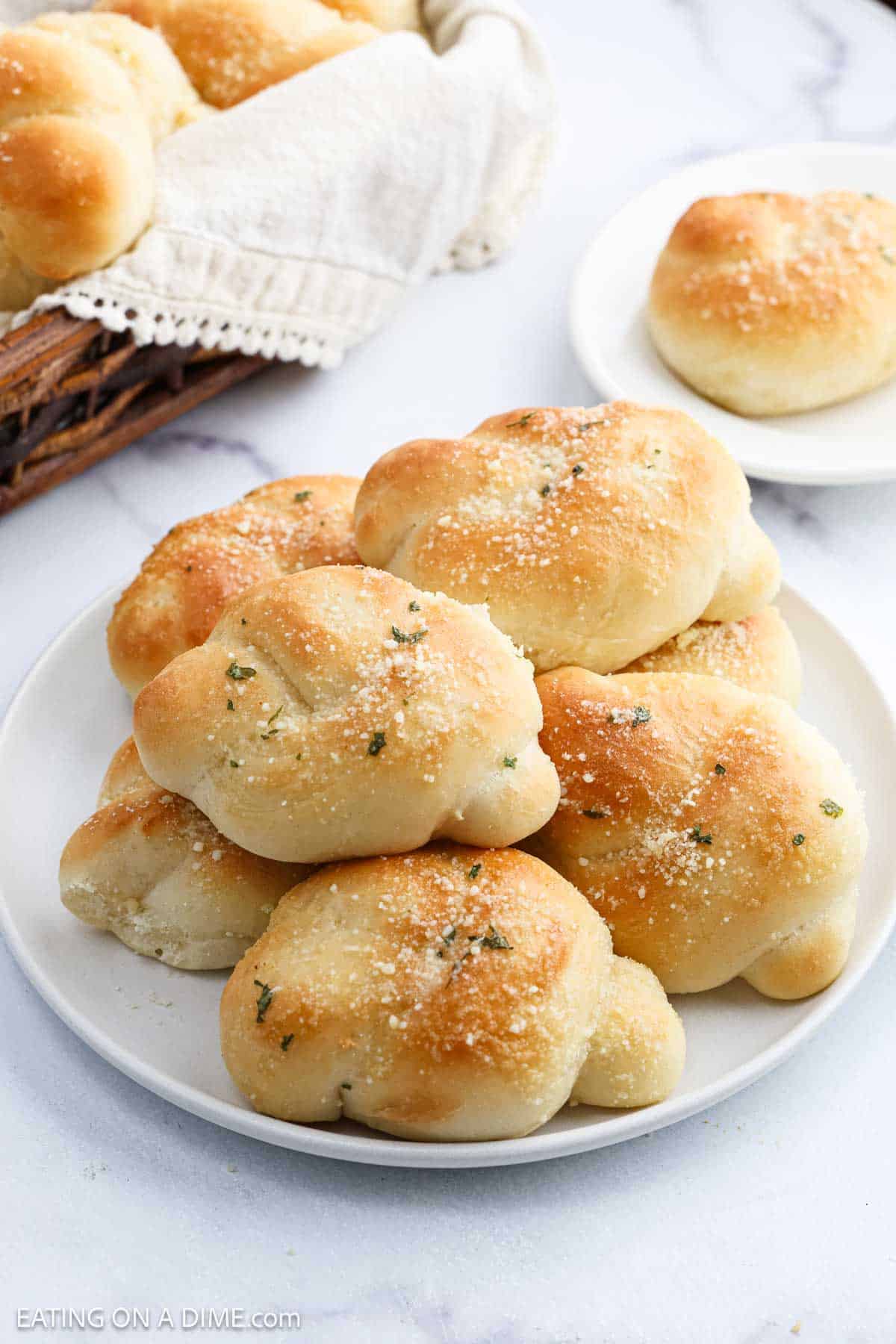 A plate of golden-brown Garlic Knots sprinkled with herbs and grated cheese sits on a white surface, with more Garlic Knots in a basket lined with a cloth in the background.
